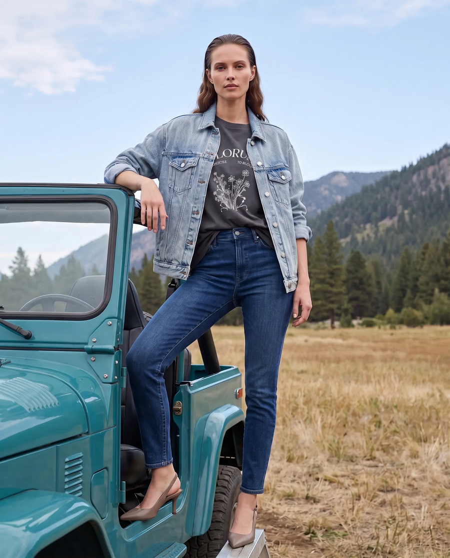 Woman in a light denim jacket and jeans leaning on a teal jeep in a mountain meadow
